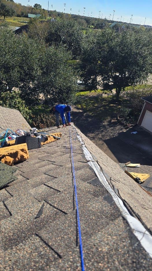 Worker installing roof shingles from a steep rooftop with tools and materials nearby.
