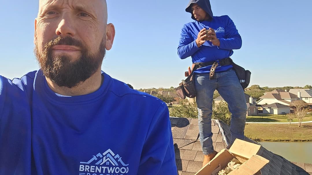 Two roofers on a rooftop, one taking a selfie while the other works with materials nearby.